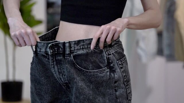 Young woman with slim waist pulls loose old jeans side to side after weight loss in home interior. Concept of wellness, proper nutrition, diet success, body transformation and clothing size reduction
