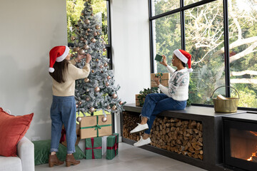 Fototapeta premium Female friends decorating flocked Christmas tree at home, wearing red Santa hats, stacking gifts