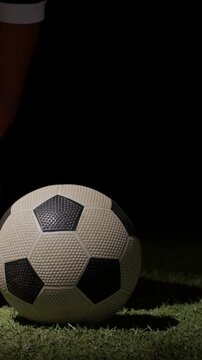 Vertical video: Walking player in socks and cleats placing ball on lit turf, steadying to kick