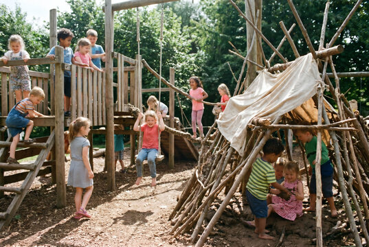 Group of diverse children playing on a modern wooden playground outdoors, climbing, sliding, and swinging in a park during summer, active childhood concept