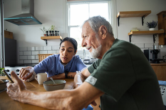 Home caregiver helps senior man use smartphone in kitchen
