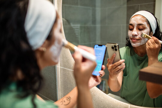 Young woman applying face mask while using smartphone in home bathroom