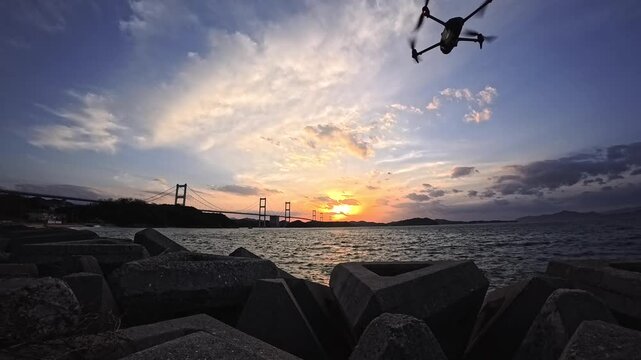 Drone flying toward Kurushima Kaikyo Bridge at sunset from Japanese coastal breakwater