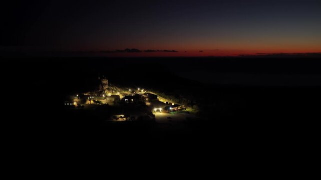 Aerial view of remote desert outpost glowing at night with red sunset horizon over dark landscape sea.