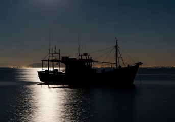 Fototapeta premium Silhouette of a fishing vessel at night reflecting on calm ocean water