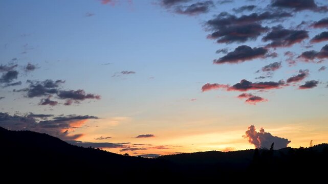 Beautiful orange sunset illuminating a colorful blue sky with soft, flowing clouds drifting over a mountain silhouette, capturing a peaceful and scenic natural landscape