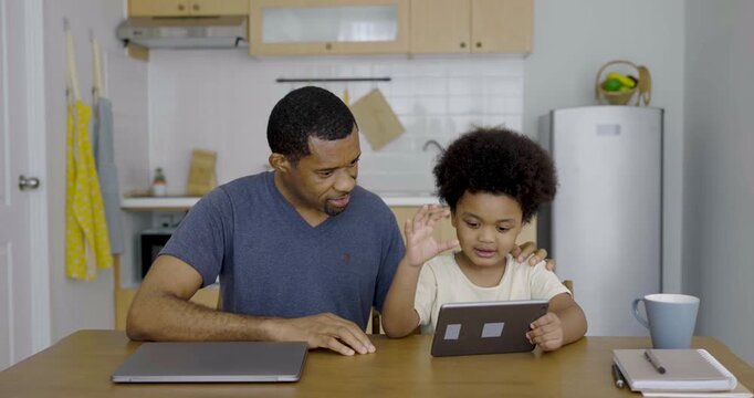 Portrait of black father sitting with young son at kitchen table while helping use a digital tablet high five while spending quality family time together
