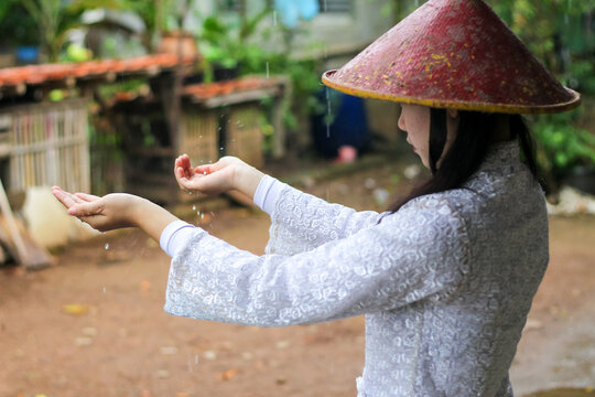 Indonesian village woman wearing traditional kebaya and conical bamboo hat standing under the rain, catching waterdrops.