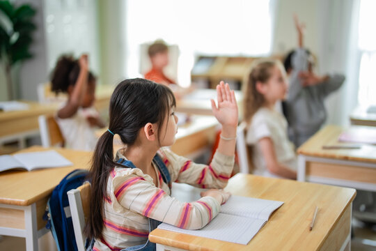 Young Asian schoolgirl raising hand while studying in elementary classroom with multicultural classmates during active learning session at school.