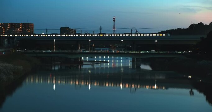 Elevated commuter train with illuminated windows moving on railway bridge reflections in calm river at night Japan. Car traffic moves on road below in blue hour. Japanese urban landscape in twilight