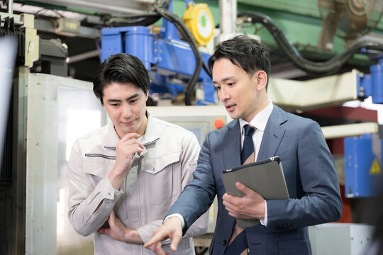 製造業の工場現場で作業着の技術者と営業が機械を見ながら見積もりや修理、メンテナンスの金額を前向きに相談する真剣に話すイメージ