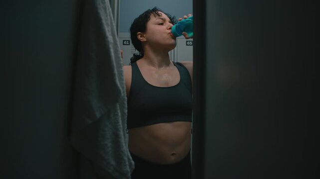 Thirsty Tired Sportswoman Drinking From Blue Bottle, Wiping Sweat on White Towel. View From Inside Locker Showing Girl Catching Breath After Fitness Session in Moody Industrial Interior. Raw Emotion.