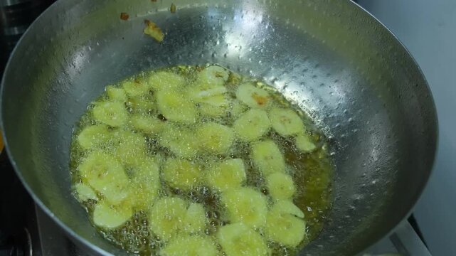 Hand dropping thin slices of green banana into a large wok of hot bubbling oil for frying chips