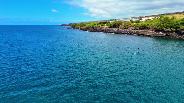 Aerial view of a person on an electric hydrofoil and a one man outrigger canoe on the turquoise ocean along the rocky coastline and lush greenery of Kawaihae, Hawaii, United States.