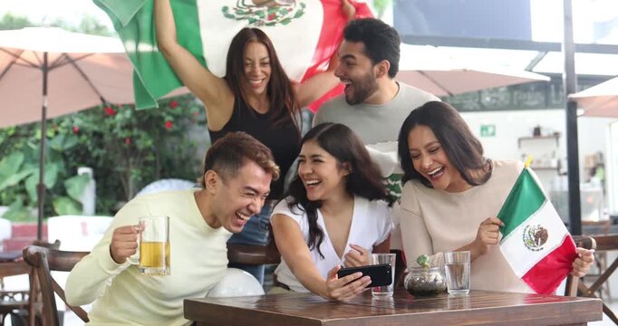 latin people soccer fans watching in phone and cheering a match game with goal, victory or winning championship in a terrace pub. Group of mexicans friends holding a flag in celebration for watching f