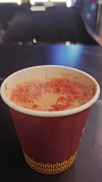 ​A close-up shot of hot, frothy fresh milk tea served in a maroon disposable paper cup on a dark surface, perfect for cafe and beverage concepts.