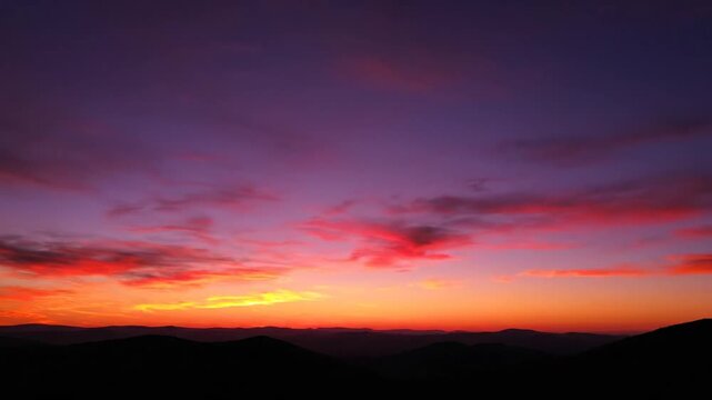 Sunset over dark mountain silhouette with vibrant gradient sky blending deep purple, orange, yellow and magenta hues featuring scattered wispy clouds during twilight golden hour.