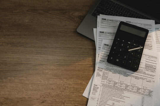 Calculator and pen placed on tax forms beside laptop on wooden desk for checking tax documents, accounting review, financial calculation, expense reporting, and tax filing workflow.