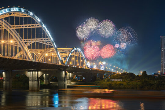 Fireworks display at Vietnam National Exhibition Center in Hanoi with Dong Tru Bridge illuminated in the foreground beside reflective river water.