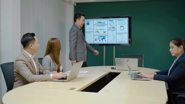 Professional Asian business team in a modern conference room. A manager joins the meeting while a female colleague distributes printed financial reports. Laptops and data charts are visible.