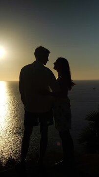 Romantic Pair Holding Hands At Dusk Near Sea. Silhouetted Lovers Share Tender Moment Facing Ocean During Sunset. Couple In Silhouette Enjoys Intimate Embrace Beside Water As Sun Sets