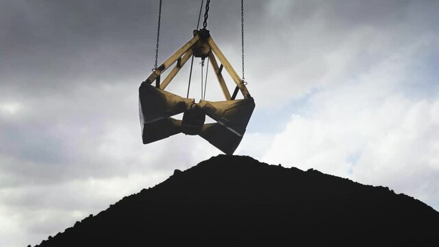Industrial clamshell grab bucket unloading coal at mining site against cloudy sky, heavy industry machinery working in open pit mine, fossil fuel extraction, raw material handling, 