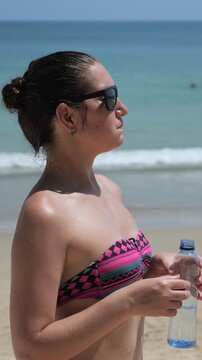 Beachside Hydration Scene. Calm Seaside Moment With Woman In Patterned Swimwear And Sunglasses. Serene Coastal Setting Showing Woman Drinking Water In Vibrant Swimwear Near Clear Ocean Waves