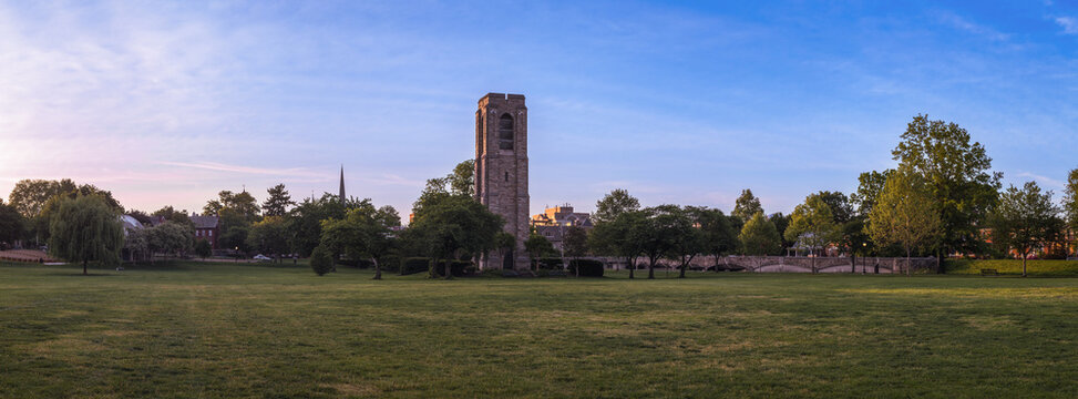 A wide panoramic view of the historic Joseph Dill Baker Carillon stone bell tower standing in a large grassy field in Baker Park, Frederick, Maryland, under a soft morning sky at dawn.