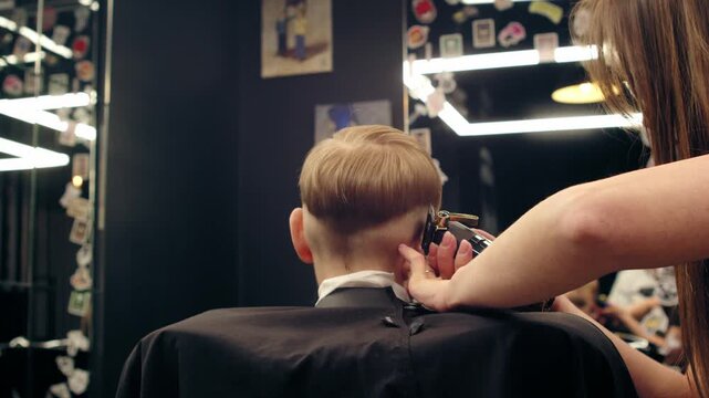 Professional female barber cutting a young boy's hair in a modern barbershop with a trimmer. Stylist trimming a child in a stylish beauty salon with a buzzer.