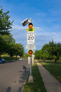 A regulatory school speed limit sign stands along Carroll Parkway in Frederick, Maryland, positioned on a concrete sidewalk next to a brick building under a bright morning sky.
