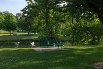 Fototapeta premium A green metal park bench sits peacefully along the grassy banks of Carroll Creek in Baker Park, Frederick, Maryland. Mature trees surround the tranquil waterside scene under a morning sky.