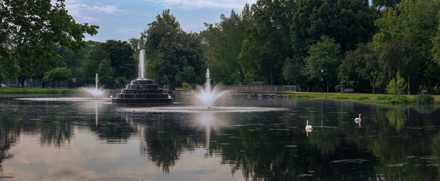 Fountains spray high and artificial swan decoys float on the calm water of Culler Lake in Baker Park, Frederick, Maryland. Green trees and a wooden footbridge line the edge under a cloudy sky.