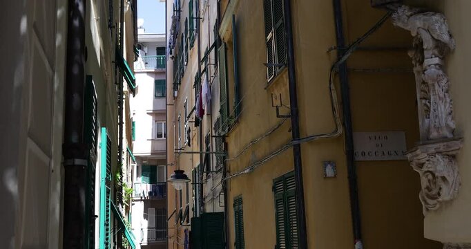 Horizontal cinematic shot of narrow Vico di Coccagna street in Genoa historic center. Panning view shows laundry drying outside windows of tall colorful Mediterranean buildings. Authentic urban scene
