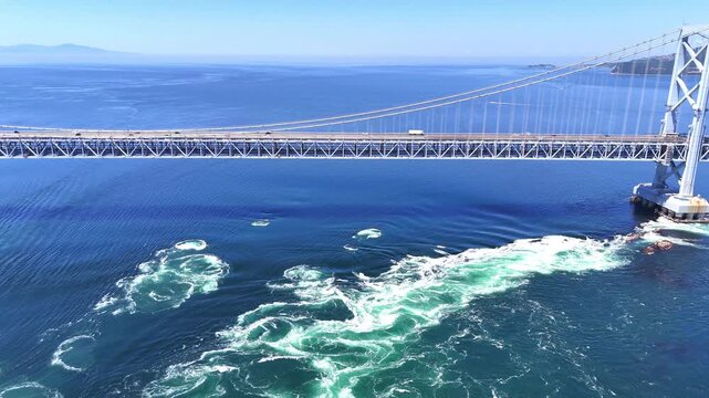 Aerial shot of the magnificent Onaruto Bridge spanning the Naruto Strait, with dynamic whirlpools and strong ocean currents visible in the foreground. A perfect blend of infrastructure and nature.
