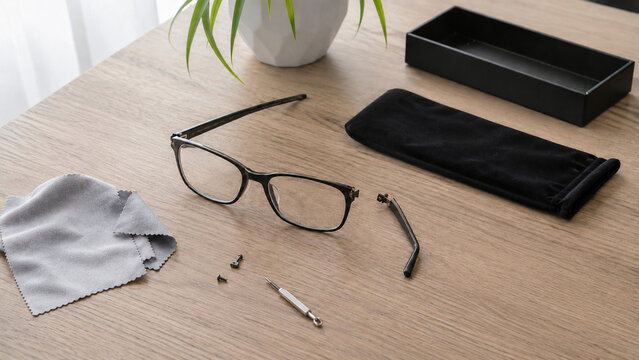 Broken black eyeglasses on a wooden desk with repair tools, cleaning cloth and case. Realistic optical service concept for vision care, eyewear maintenance and professional repair.