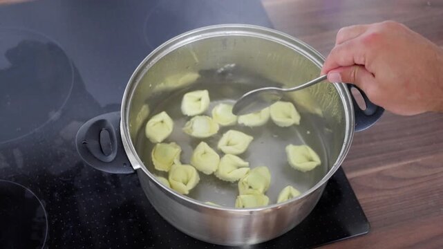 Stirring Stuffed Pasta On Stove. Closeup Of Stirring Yellow Tortellini In Stainless Pot With Steam Rising. Preparation Scene Showing Chef Inspecting Cooked Stuffed Pasta On Induction Stove Top