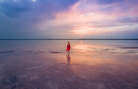 Aerial of a young woman in red dress walking in the water of a unique pink salt lake. Sunset at lake Bursol with beautiful reflections on calm water surface. Stunning scenery
