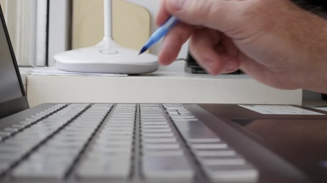 Hand Holding Blue Pen Writing on Laptop Keyboard. Typing on Laptop Keyboard Closeup Detail.