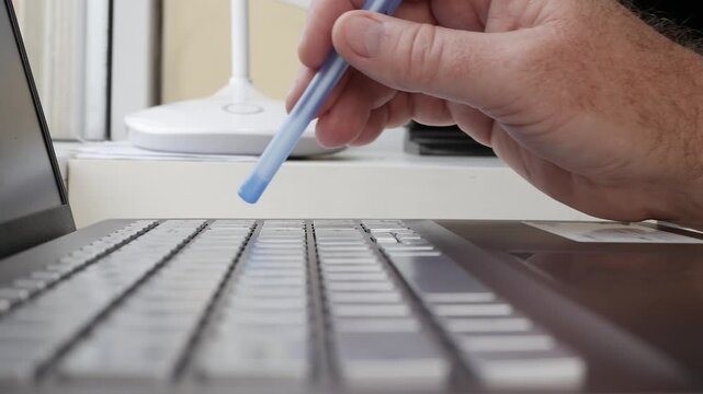 Hand Holding Blue Pen Writing on Laptop Keyboard. Typing on Laptop Keyboard Closeup Detail.