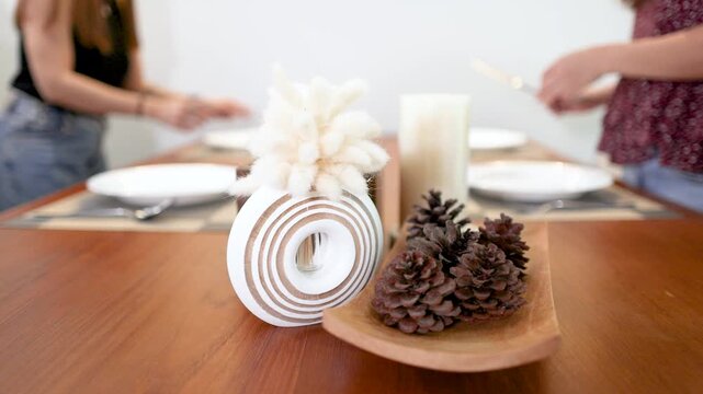 Two Women Setting a Minimalist Dining Table with Pine Cone Centerpiece