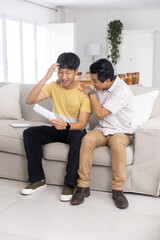 Obraz premium Asian father and son sitting on light-gray couch in living room, holding white envelope and tablet
