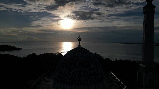 Aerial view of the Masjid Raya Nur Ilahi dome silhouetted against a golden sunset over the sea in Bukit Bestari, Riau Islands, Indonesia.