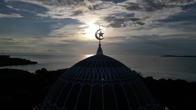 Aerial view of the dome of Masjid Raya Nur Ilahi with its crescent and star symbol silhouetted against the sunset over the sea in Bukit Bestari, Riau Islands, Indonesia.