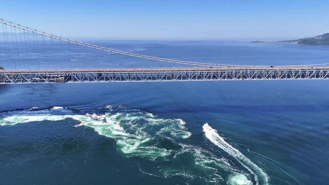 Dynamic 4K aerial footage showing a boat navigating the powerful currents of the Naruto Strait directly beneath the massive steel structure of the Onaruto Bridge. Excellent representation of logistics