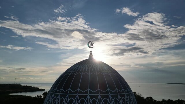 Aerial view of the dome of Masjid Raya Nur Ilahi with its crescent and star symbol against a bright sky over the sea in Bukit Bestari, Riau Islands, Indonesia.