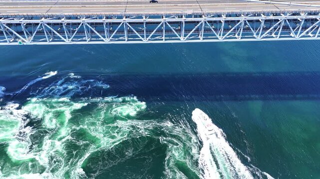 A visually striking 4K aerial view looking down at the Onaruto Bridge and the vibrant emerald green waters of the Naruto Strait. A boat cuts through the dynamic whirlpools and tidal currents, creating