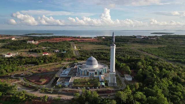 Aerial view of Masjid Raya Nur Ilahi with its blue dome and tall minaret overlooking the coast and lush greenery in Bukit Bestari, Riau Islands, Indonesia.