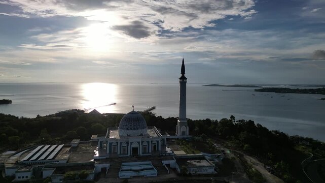 Aerial view of Masjid Raya Nur Ilahi with its grand dome and tall minaret overlooking the calm sea under a bright sun Bukit Bestari, Riau Islands, Indonesia.