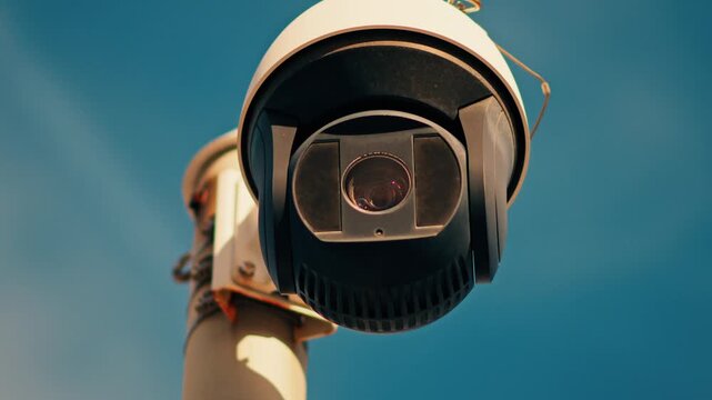 Modern dome security surveillance camera rotating on metal pole against clear blue sky.