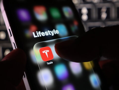 Closeup of a persons finger touching the tesla app icon on a smartphone screen, representing electric vehicle technology and digital lifestyle